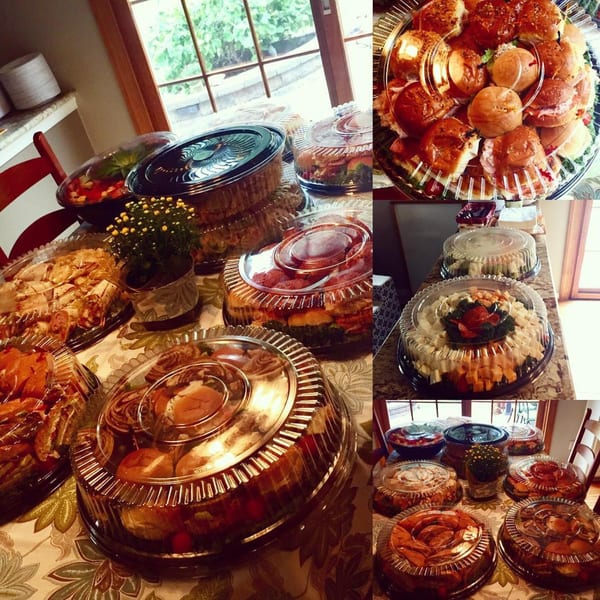 Multiple catering trays of Italian food, including sandwiches, salads, and appetizers, on a patterned tablecloth.